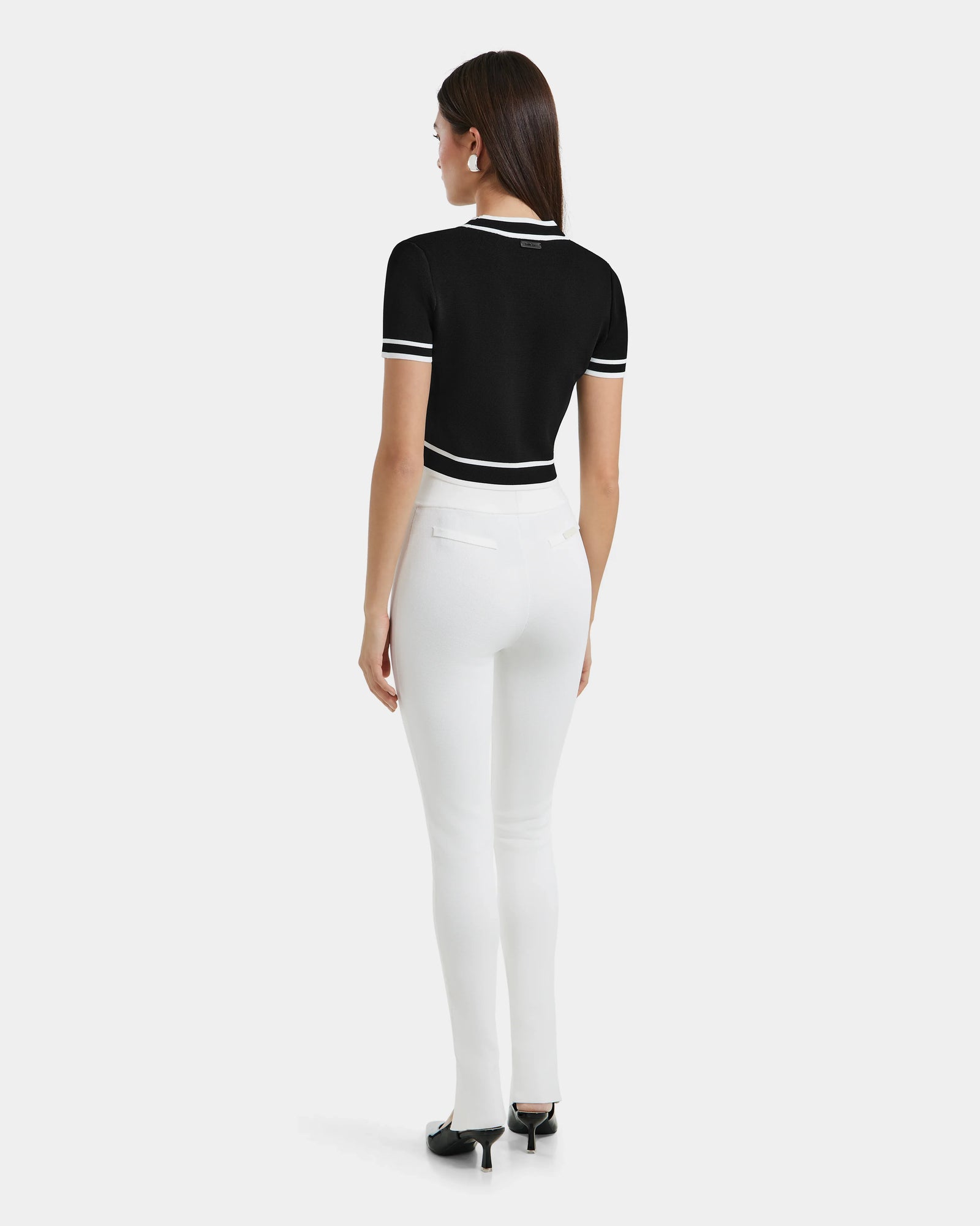 A woman faces away, wearing a short-sleeve black top with white trim and fitted white MATHILDA - 8524515 Iceland leggings by Rudsak with front ankle slits, paired with black heels against a plain light background.
