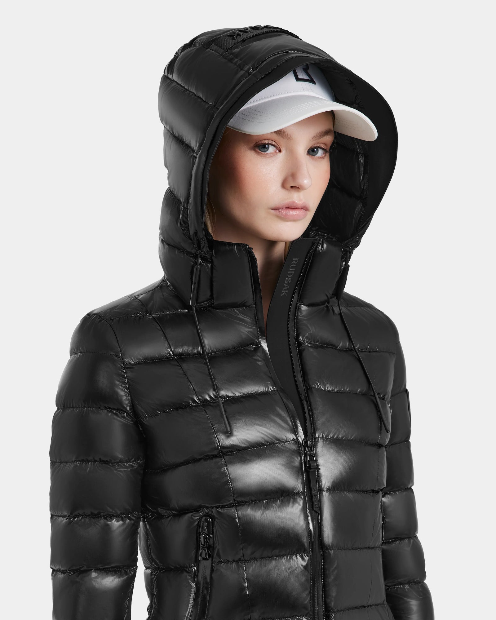 A woman wearing the MELISSA - 8121000 BLACK shiny black puffer jacket with a hood and a white baseball cap looks slightly to the side against a plain light background.