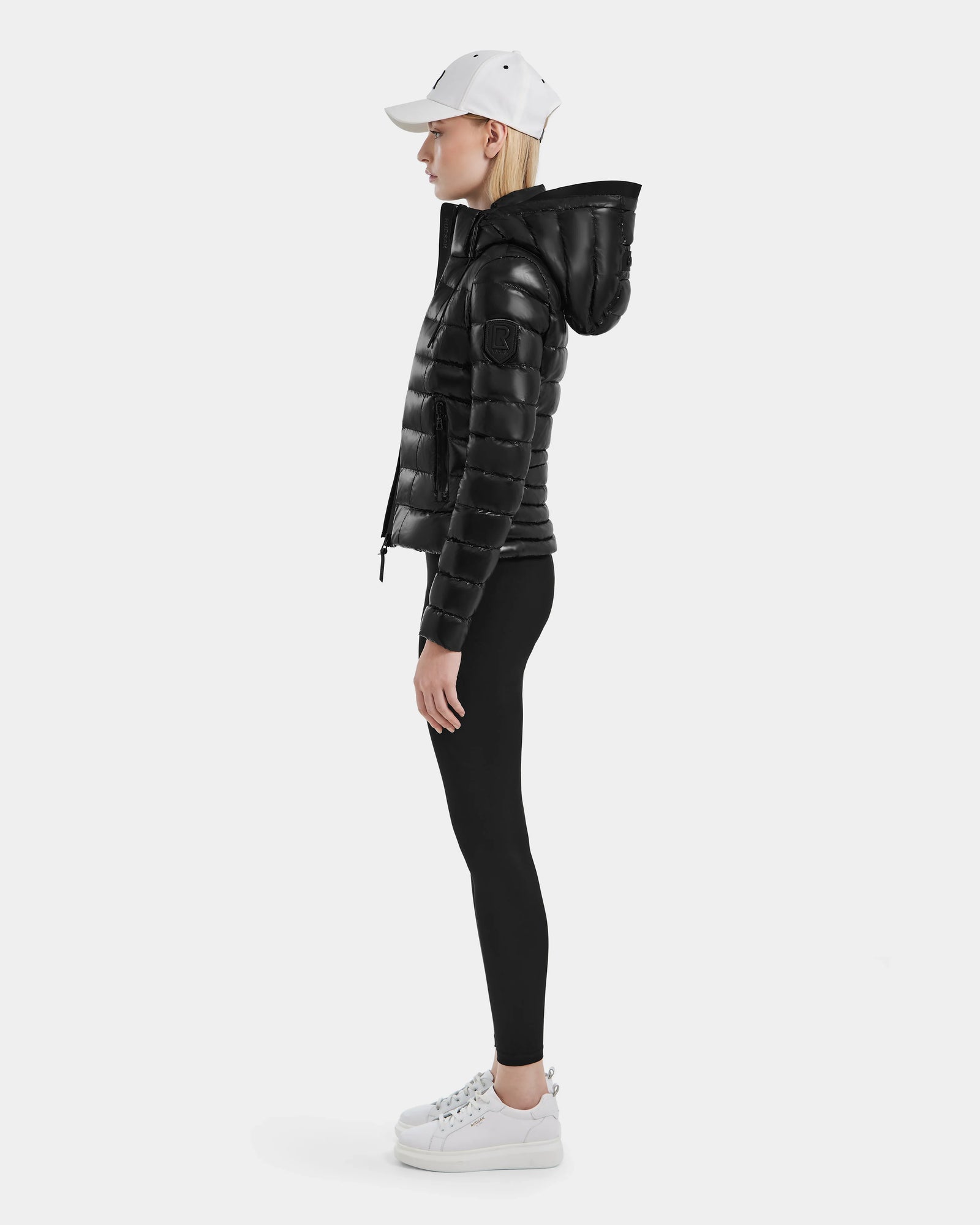 A woman in a white baseball cap, RDS certified down puffer jacket, black leggings, and MELISSA - 8121000 BLACK sneakers stands in profile against a plain white background.