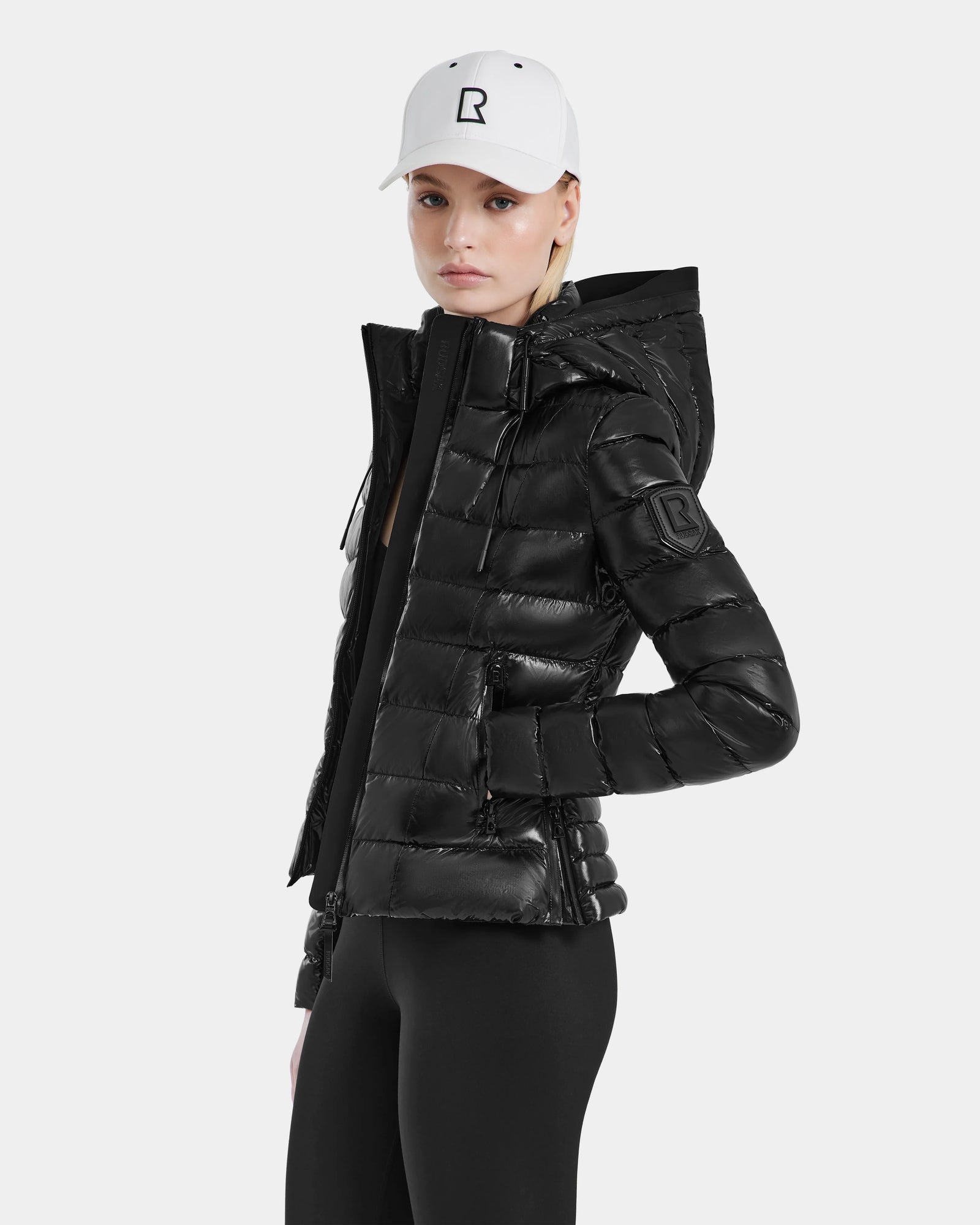 Wearing the Excluded MELISSA - 8121000 BLACK fitted puffer jacket, black leggings, and a white baseball cap with an R logo, a person stands against a plain light background.