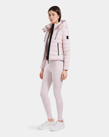 A woman with straight dark hair and a neutral expression poses against a plain background, wearing the Rudsak MELISSA - 8125054 Light Pink puffer jacket, matching leggings, a white top, and white sneakers.