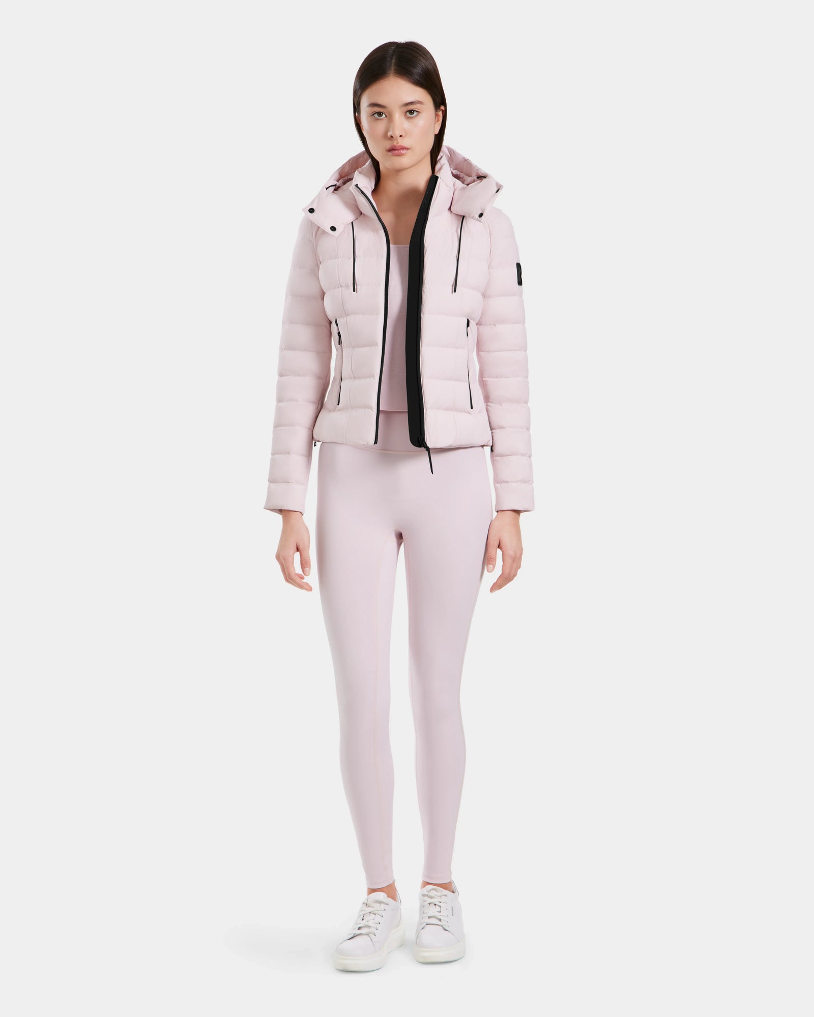 A woman faces forward, wearing the MELISSA - 8125054 Light Pink water-resistant puffer jacket by Rudsak with a hood, matching pink leggings, and white sneakers against a plain white background.