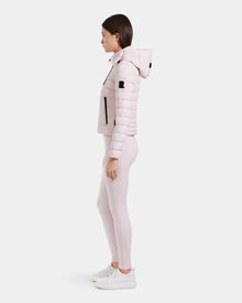 A woman stands in profile wearing the Rudsak MELISSA - 8125054 light pink puffer jacket, a water-resistant, lightweight down coat with a hood, paired with matching light pink leggings and white sneakers against a white background.