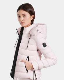 A young woman models the Rudsak MELISSA - 8125054 Light Pink puffer jacket, featuring a hood, black zipper accents, and a signature “R” patch on the sleeve—highlighting this stylish, water-resistant outerwear against a plain background.