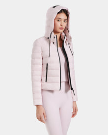 A woman in the Rudsak MELISSA - 8125054 Light Pink water-resistant puffer jacket with a hood, paired with matching light pink leggings and top, stands turned slightly to the side against a white background.