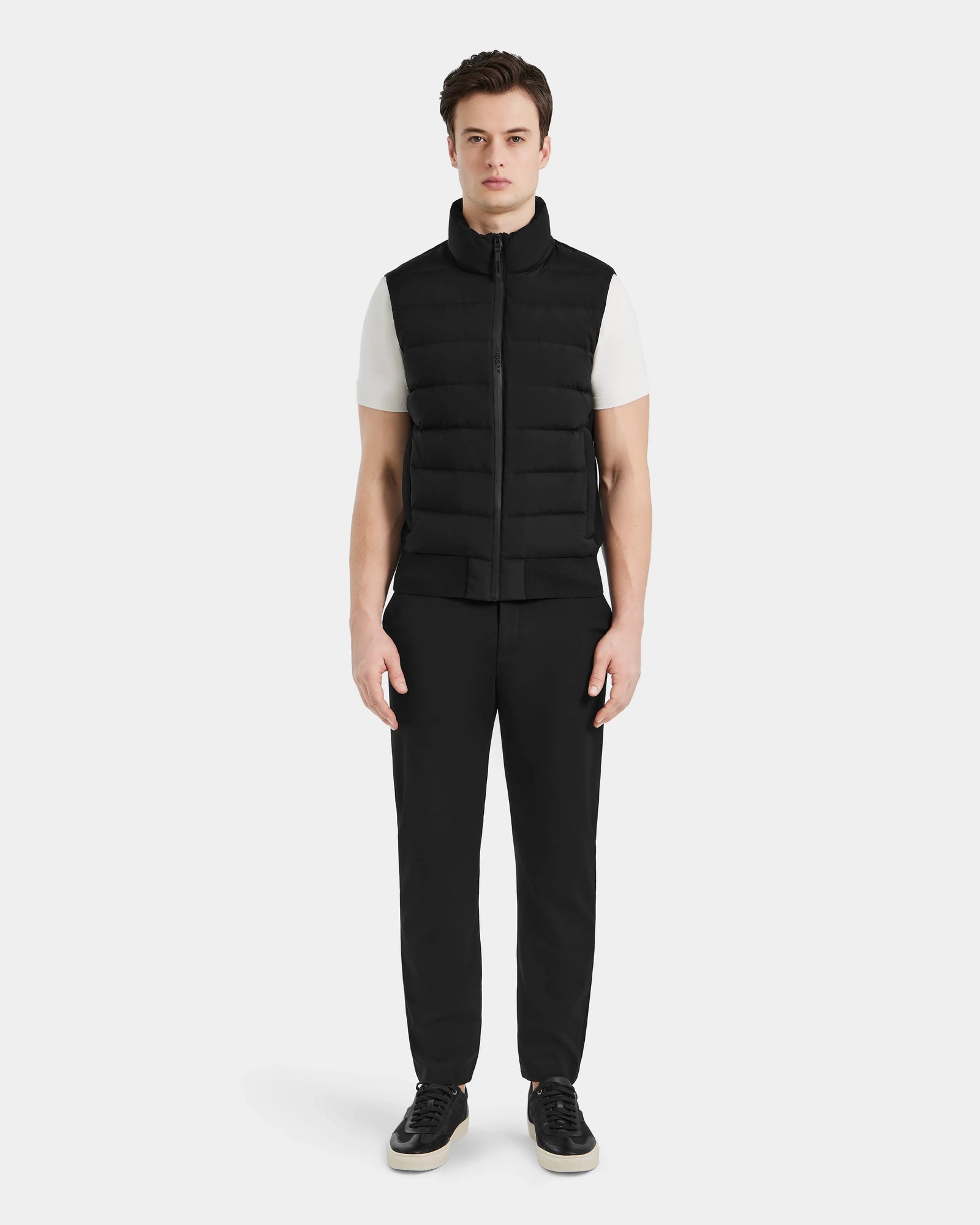 A man faces forward against a plain white background, wearing the Rudsak MERLO - 6124017 Black quilted vest over a white short-sleeve shirt, paired with black pants and black sneakers with white soles.