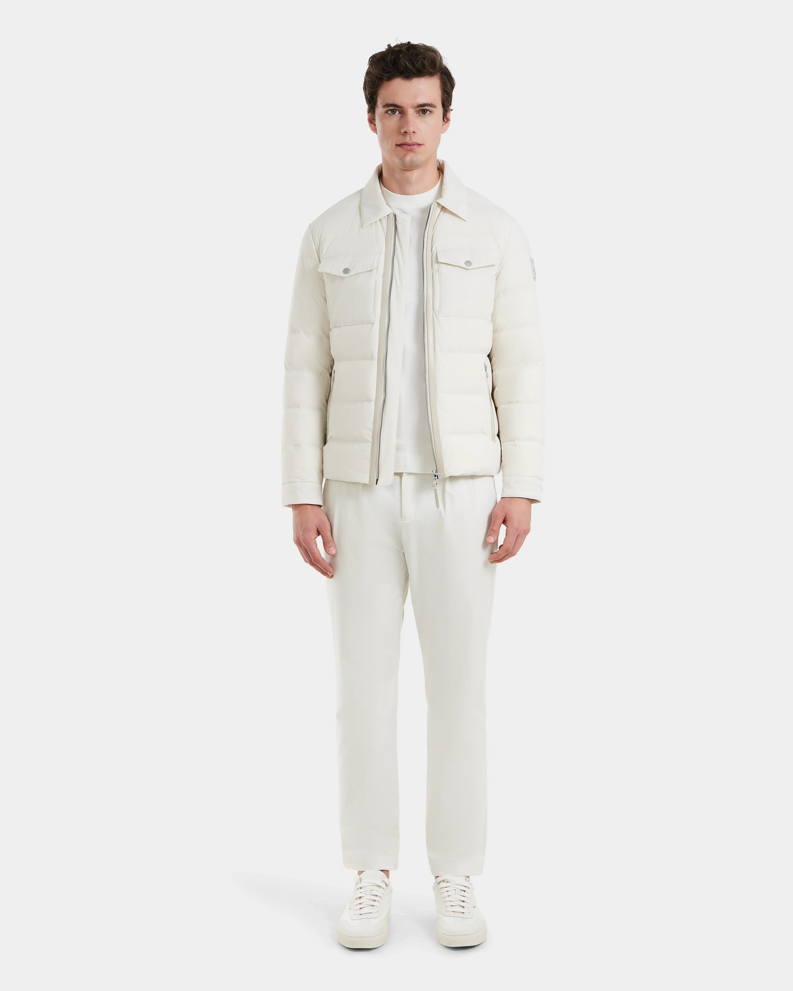 A man stands facing forward in a plain white setting, wearing the Rudsak ORNO - 6124601 Vanilla: a lightweight insulated jacket, t-shirt, drawstring pants, and sneakers.