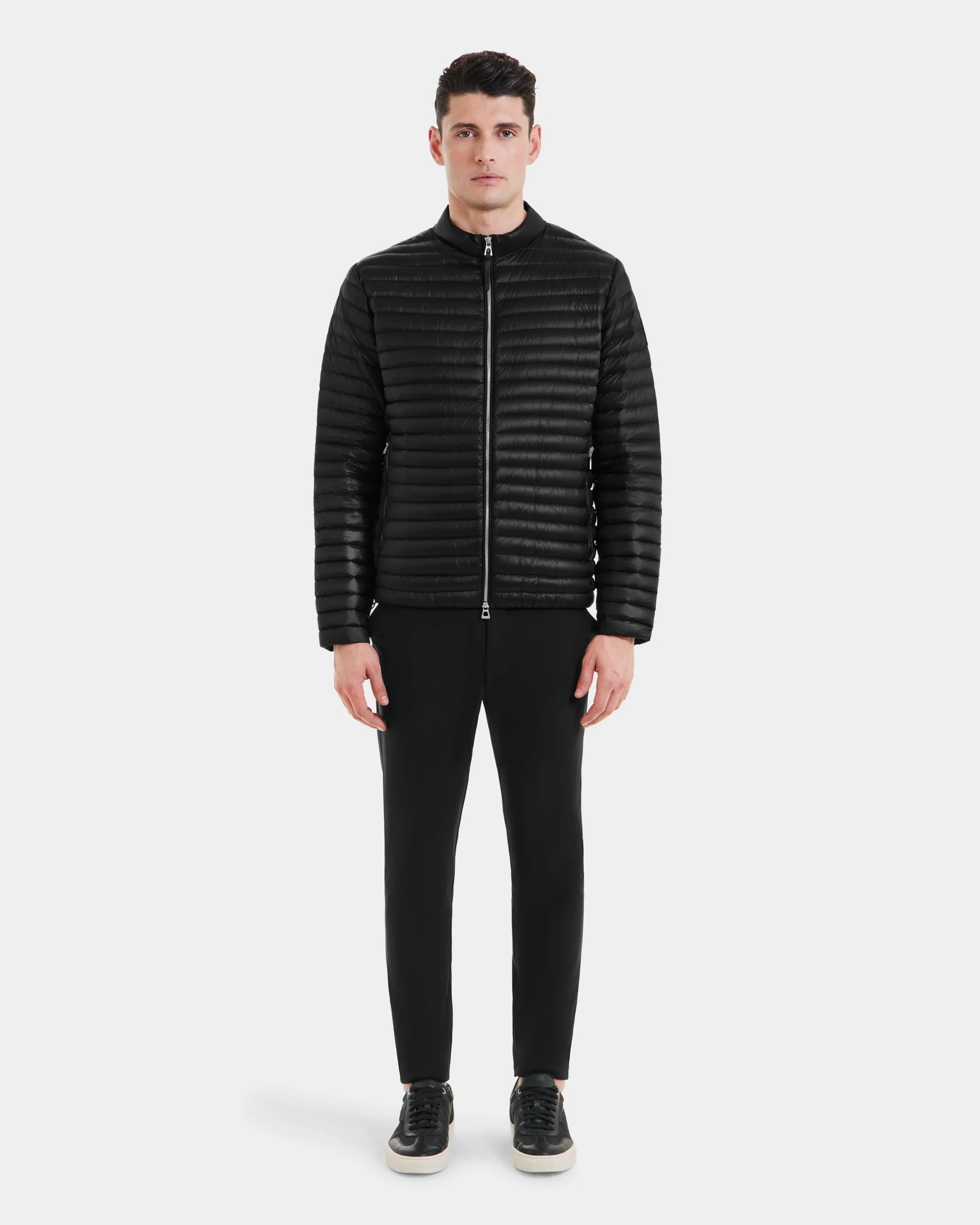 A man stands facing forward against a plain background, wearing the REYMOND - M125013 Black men’s down jacket by Rudsak, made from recycled nylon with RDS-certified down, paired with black pants and black sneakers with white soles.