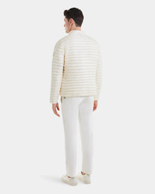 A man stands facing away in the Rudsak REYMOND - M125013 Iceland, a cream-colored quilted men’s down jacket made from recycled nylon, paired with white pants and sneakers against a plain light background.