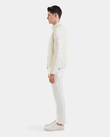 A man stands in profile wearing the REYMOND - M125013 Iceland off-white quilted men’s down jacket by Rudsak, filled with RDS-certified down, paired with white pants and sneakers against a plain white background.