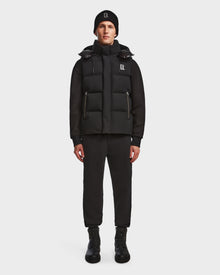 A man stands facing forward against a plain white background, wearing the Rudsak REN M - 9123512 Black M weather-resistant puffer vest with RDS-certified down over a hoodie, paired with jogger pants, lace-up boots, and a logo beanie.