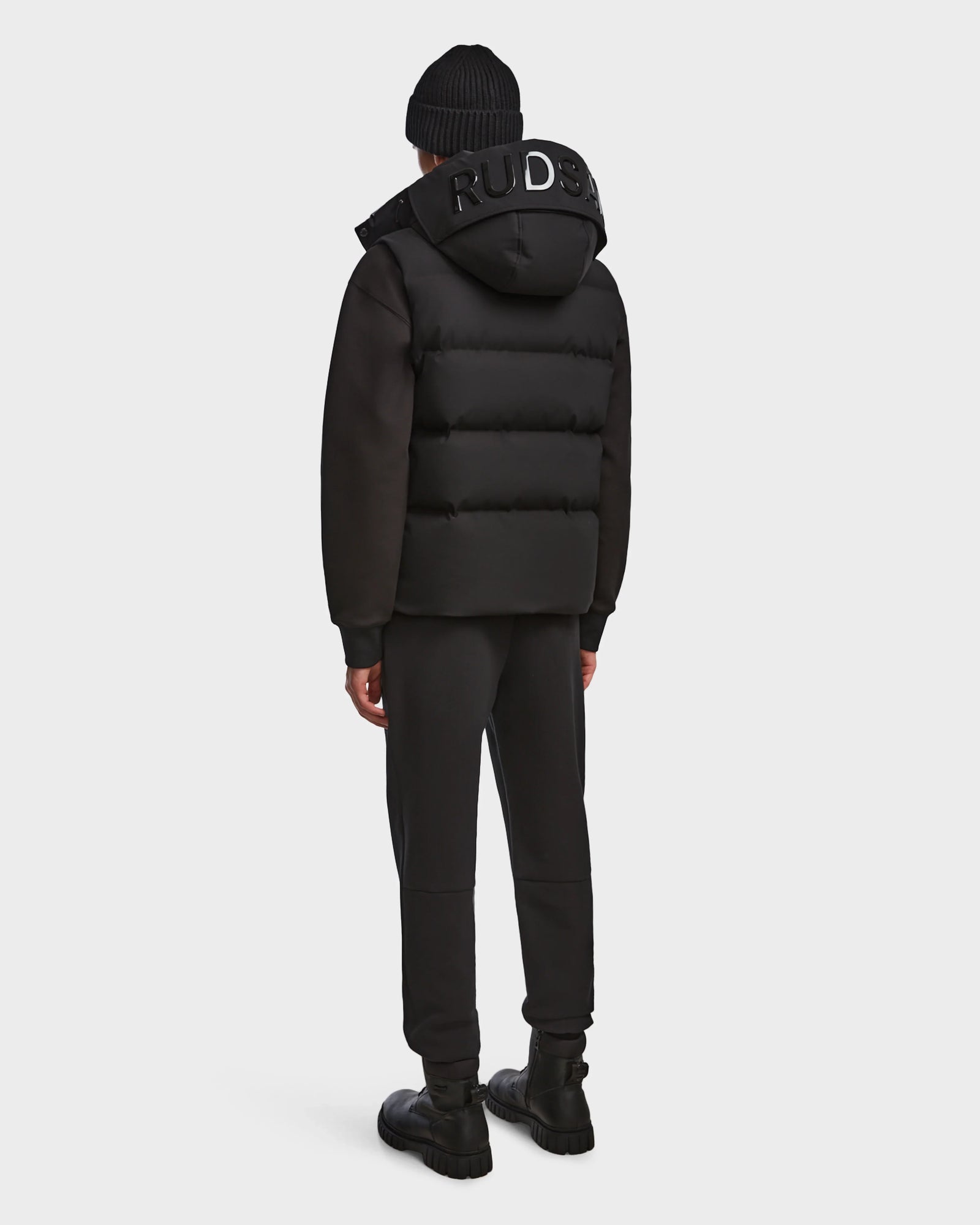 A person in the Rudsak REN M - 9123512 Black M weather-resistant puffer jacket, black beanie, pants, and shoes stands facing away from the camera against a plain light background.