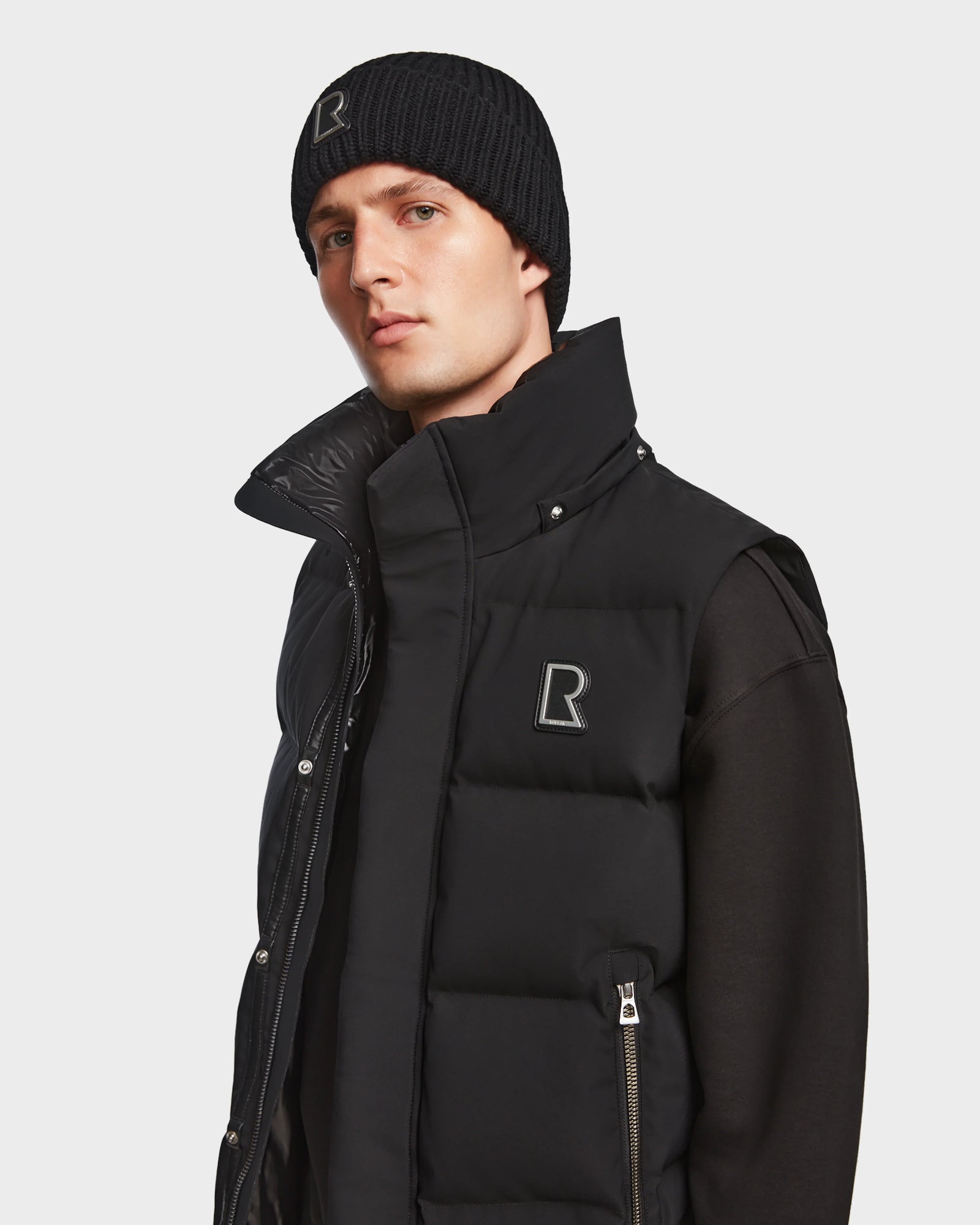 A man models the Rudsak REN M - 9123512 Black puffer vest with an R logo, paired with a black long-sleeve shirt and black Rudsak beanie, against a plain light background.