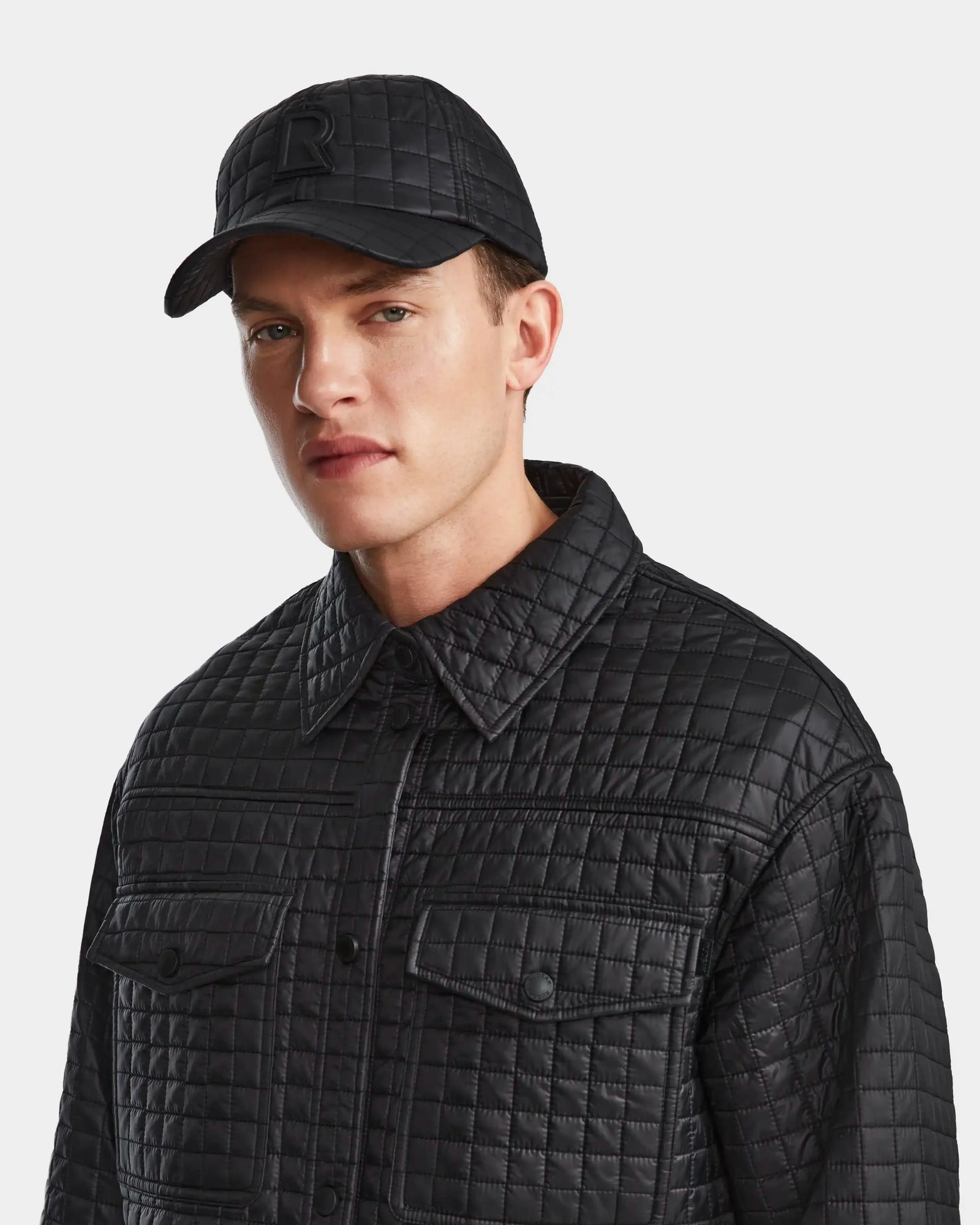 A man models the Rudsak KERRY - 9124003 Black M quilted shirt jacket with eco-friendly insulation and large front pockets, paired with a matching black quilted cap, against a plain light backdrop.