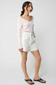 Woman wearing a light pink cardigan and white shorts on a plain background