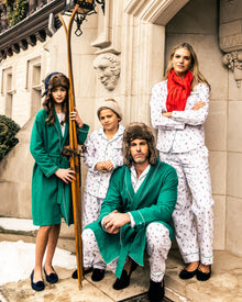 A family in festive winter attire poses by an ornate building. The man and two kids wear fur hats, the woman a red scarf. The girl holds wooden skis with snow on the ground. The childrens sleepwear features Petite Plumes Kids Flannel Pajama Set in Apres Ski for cozy warmth.