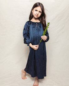 A young girl with long brown hair stands barefoot against a neutral background, wearing a Petite Plume Girls Silk Delphine Nightgown in Navy Polka Dot. She holds a green leafy branch, exuding innocence and charm.