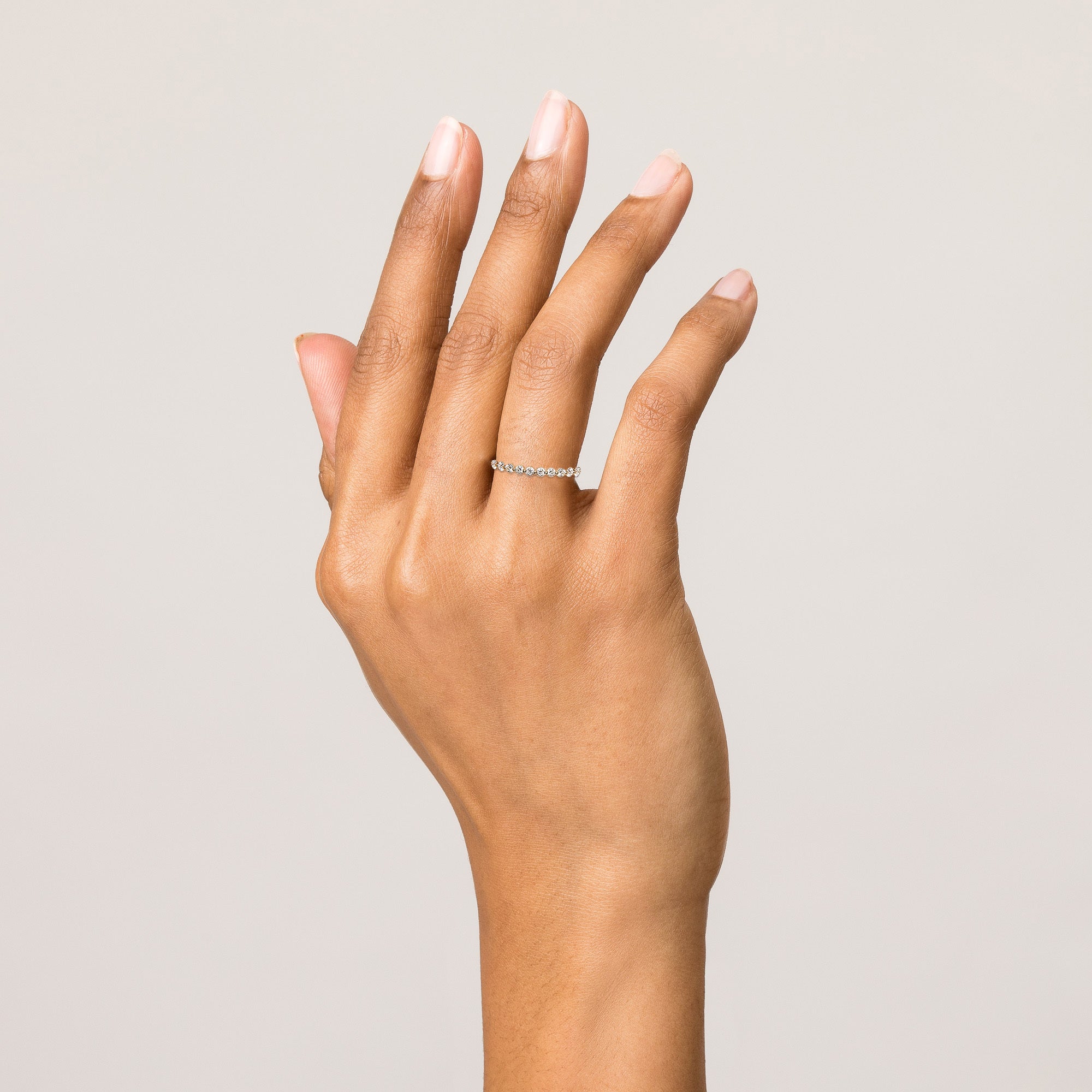 Model Close-up of a woman's hand raised gracefully with gently spread fingers against a smooth light background. On her ring finger, she is wearing a single delicate gold band studded with a row of small diamonds.