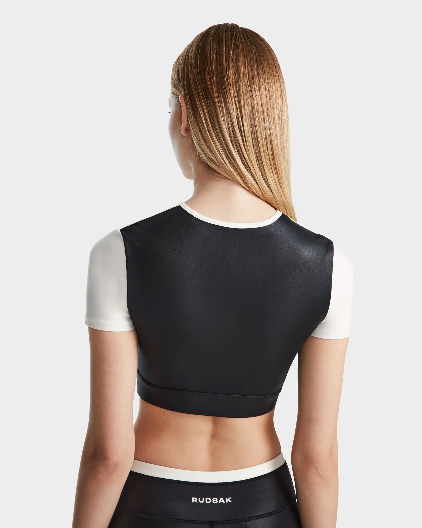 A woman with straight, blonde hair is shown from the back, wearing the LILIANE - 8524018 Black crop top by Rudsak with white sleeves and matching black bottoms featuring RUDSAK on the waistband.