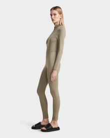 A woman with straight blonde hair stands sideways, wearing the Rudsak SHANYA - 8523526 Cement Pattern fitted long-sleeve zip-up bodysuit and black slide sandals against a plain white background.
