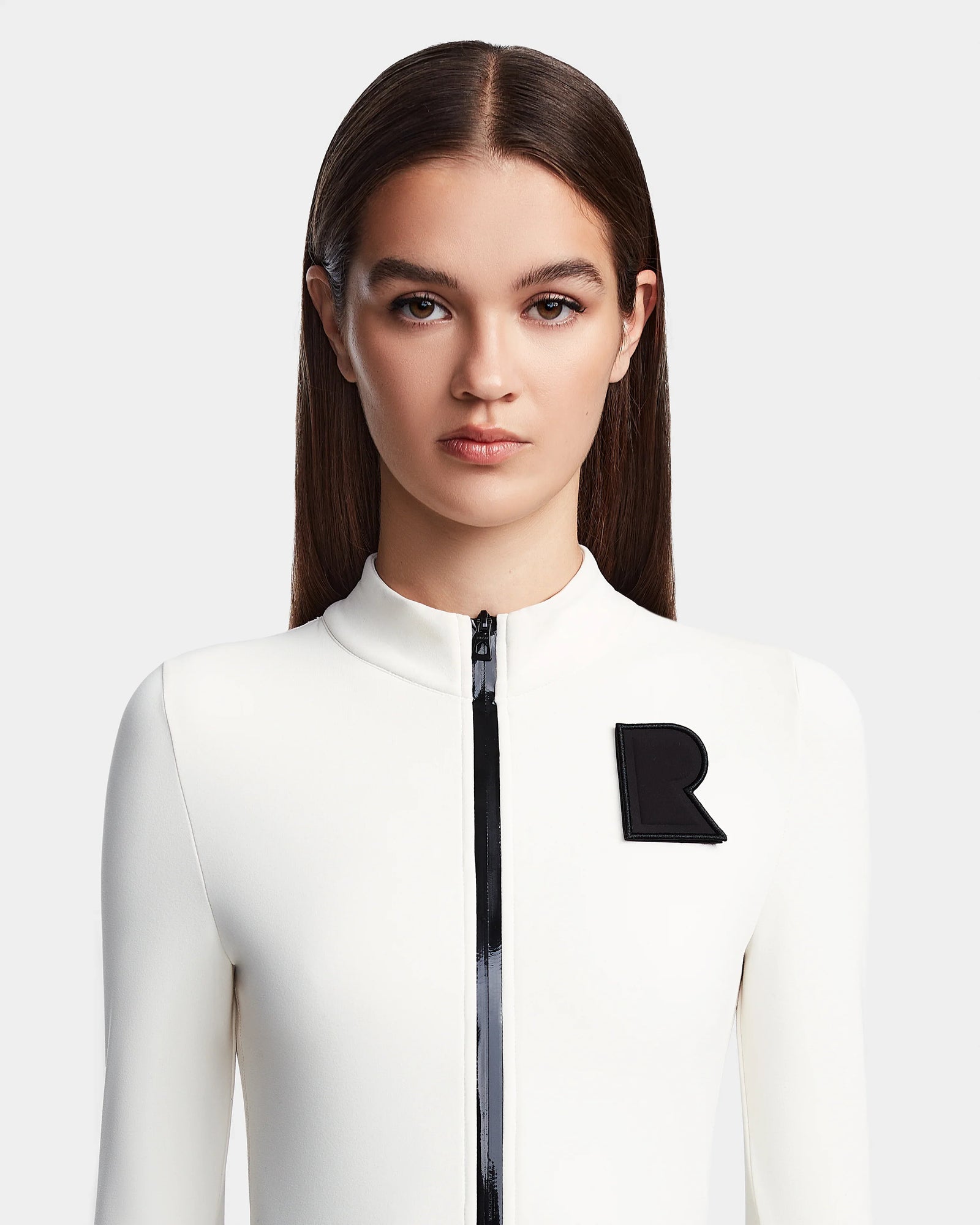 A young woman with straight brown hair models the Rudsak DAVINA - 8523534 Iceland: a fitted, fleece-lined bodysuit with long sleeves, black zipper, and bold black R patch, evoking après-ski style against a plain light grey background.