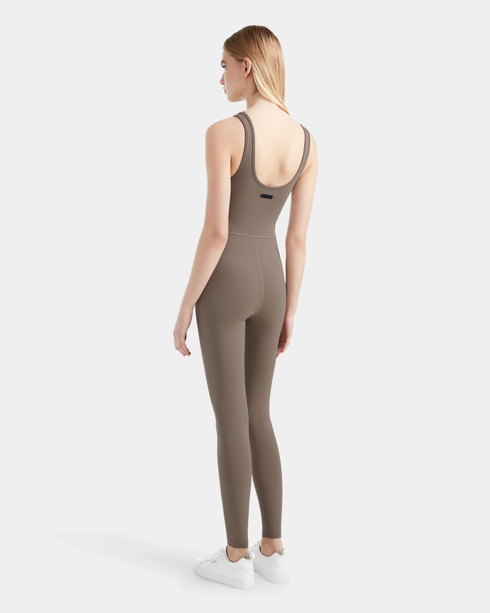 A woman faces away, wearing the Rudsak JEWEL - 8524032 Cement jumpsuit, featuring a fitted stretch tank design, paired with white sneakers against a plain light gray backdrop.