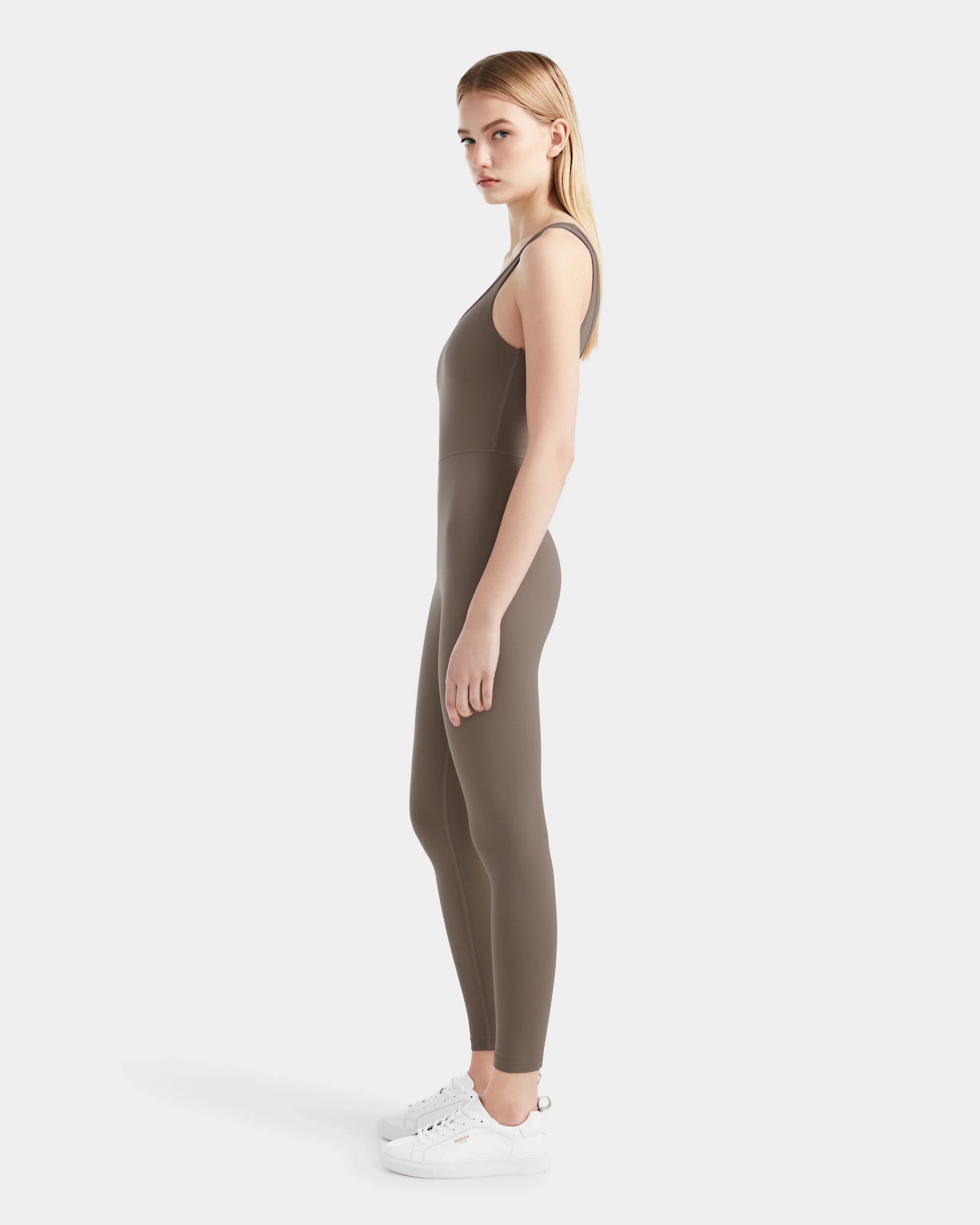 A woman in profile wears the Rudsak JEWEL - 8524032 Cement fitted, sleeveless jumpsuit made from stretch fabric with white sneakers, set against a plain white background.