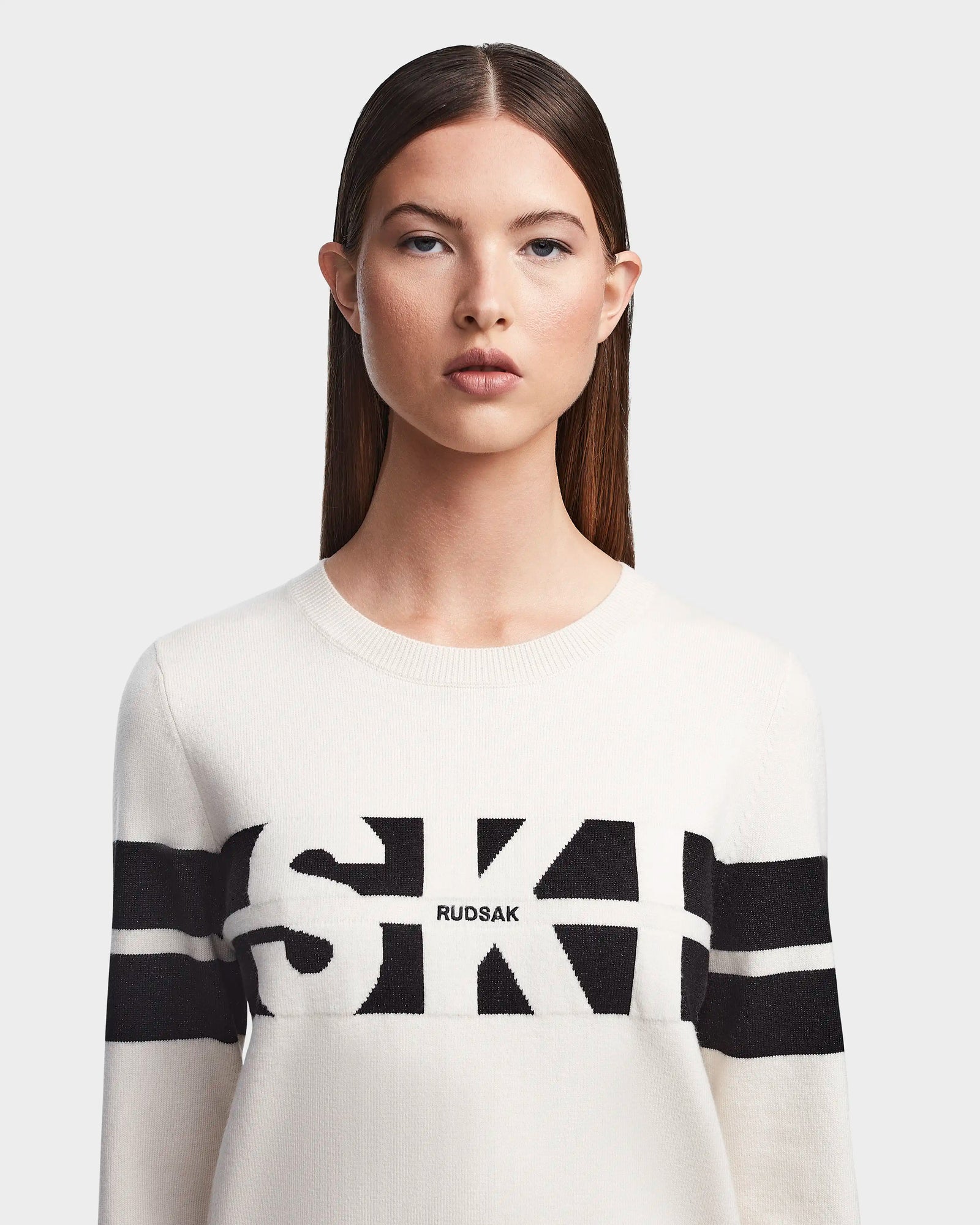 A woman with straight brown hair and a neutral expression wears the Rudsak ODESSA - 8523540 Iceland après-ski sweater, featuring soft white knit with black horizontal stripes and RUDSAK printed across the chest, against a light gray background.