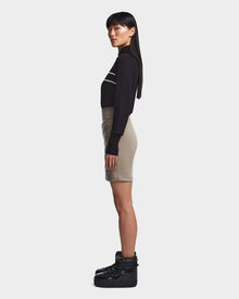 A woman stands in profile against a plain background, wearing the Rudsak MARZI - 8523528 Cement high-waisted beige winter skirt in glossy fabric, paired with a black long-sleeve top with white stripes and black platform boots.