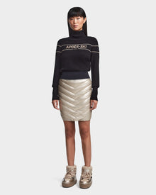 A woman faces forward against a white background, wearing Rudsaks MARZI - 8523528 Metallic Ivory gold chevron quilted winter skirt and matching gold padded boots, styled with a black APRES-SKI turtleneck sweater.