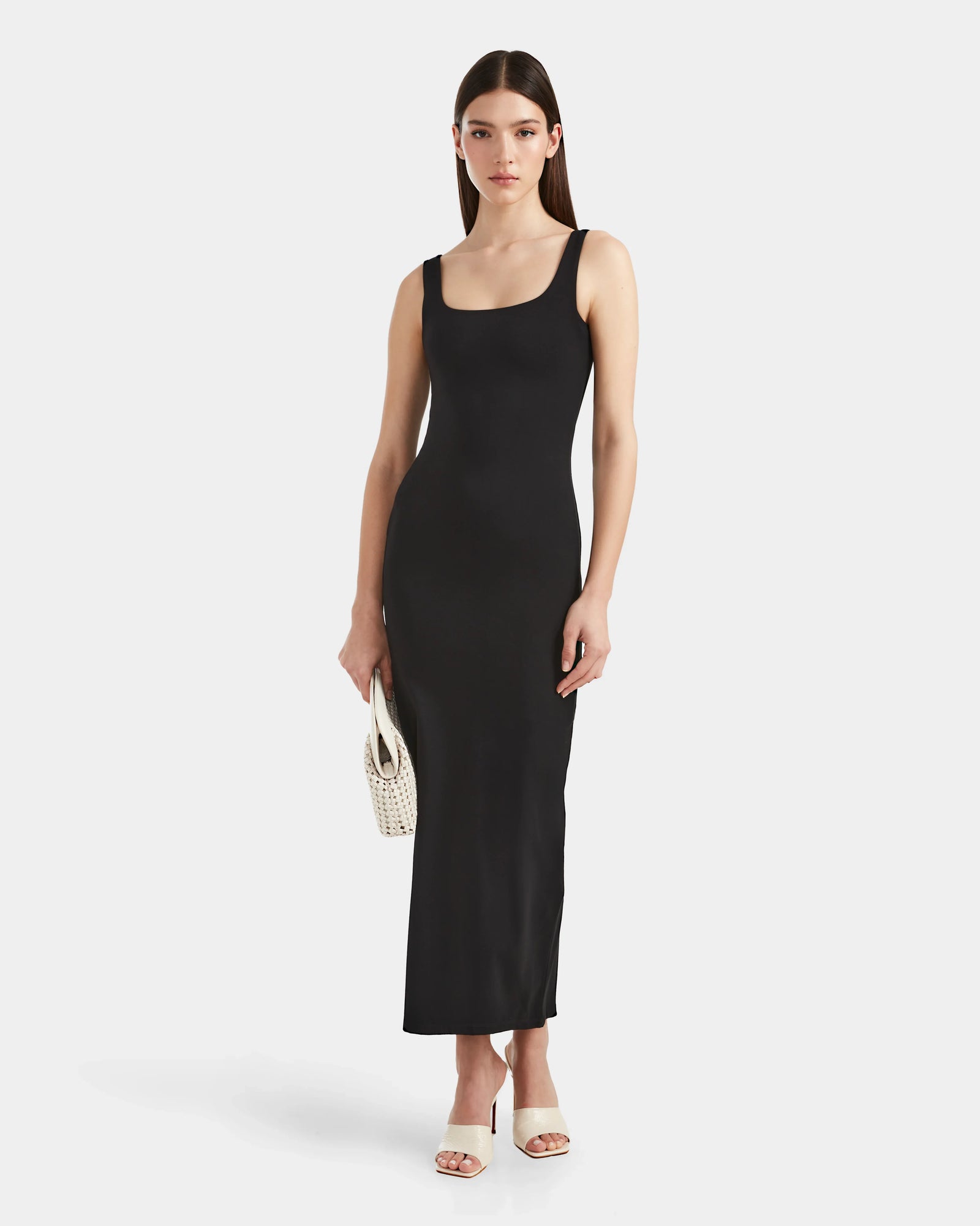 A woman wears the Rudsak WANDA - 8524035 Black fitted sleeveless maxi dress with stretch performance fabric, paired with white high-heeled sandals and a small white woven handbag, against a plain white backdrop.