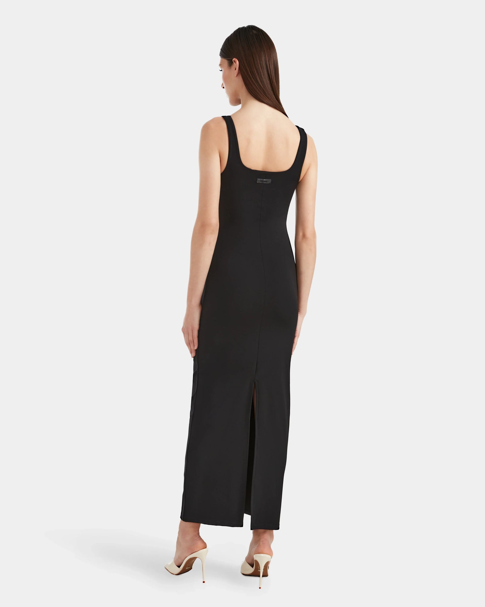 A woman stands facing away, exuding elegance in the Rudsak WANDA - 8524035 Black fitted, sleeveless maxi dress with a scoop back and hem slit, paired with white high heels.