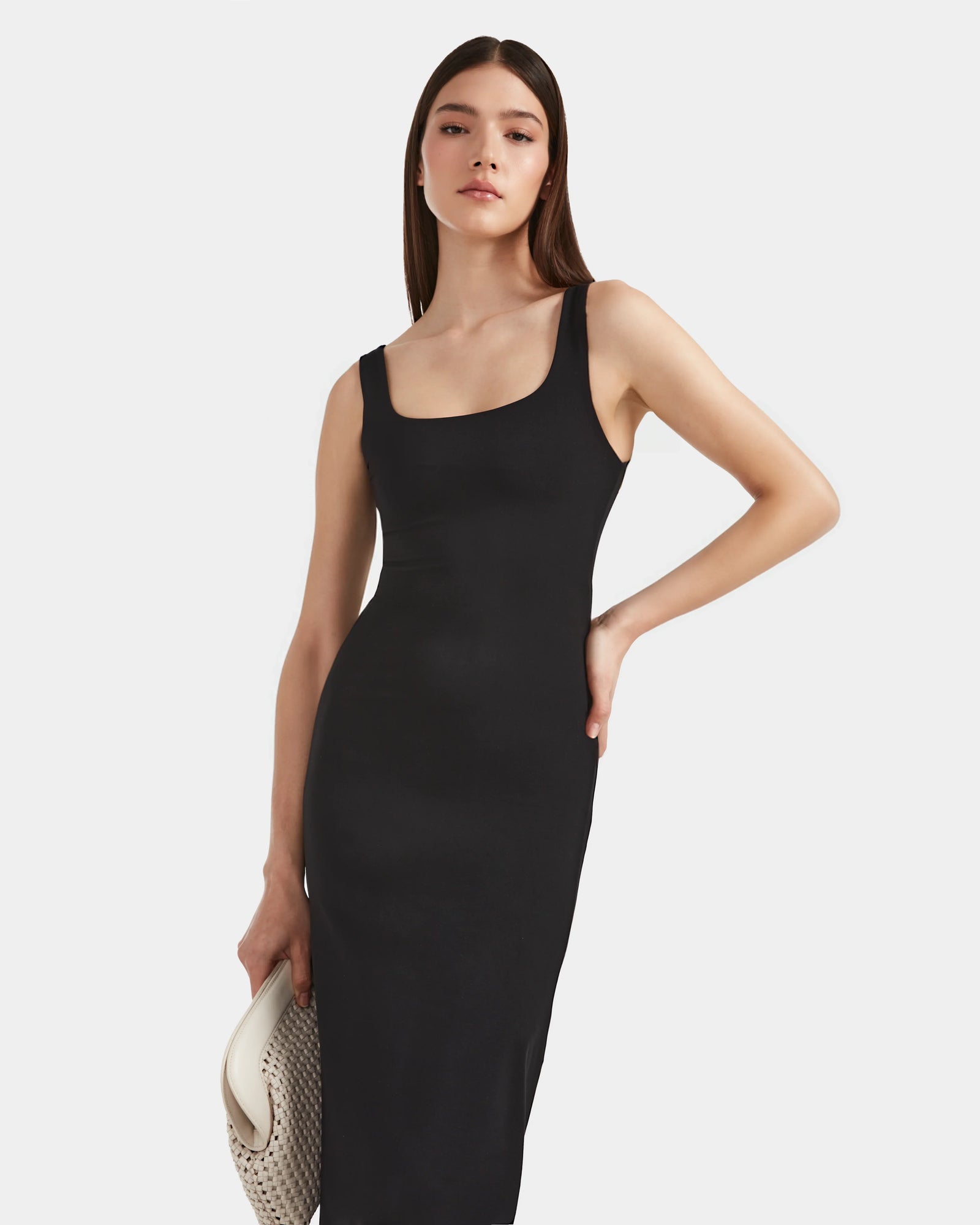 A woman with straight brown hair wears the Rudsak WANDA - 8524035 Black sleeveless fitted dress made from stretch performance fabric, holding a white woven clutch against a plain white background, exuding contemporary elegance.