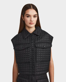 A woman with long brown hair wears the Rudsak SOLSA - 8124048 womens sleeveless black cropped quilted vest with a collar and front pockets, standing against a plain white background.