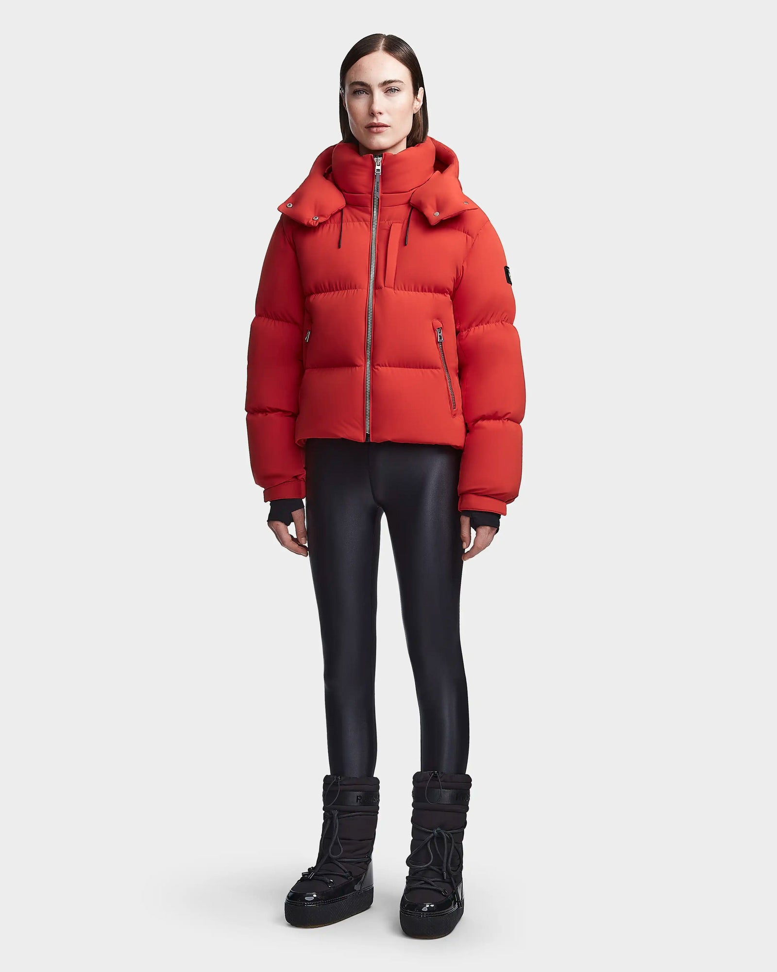 A woman faces forward, wearing the Rudsak MAVE M - 8123572 Orange womens down puffer, shiny black leggings, and black winter boots against a plain light gray background.