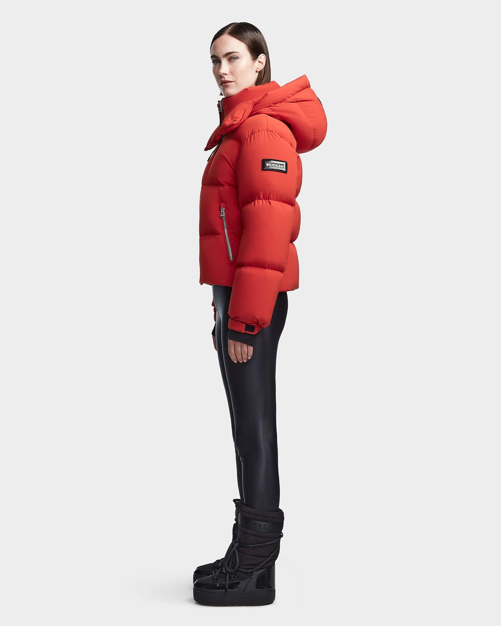 A woman stands in profile wearing the Rudsak MAVE M - 8123572 Orange down puffer jacket with a hood, paired with black shiny leggings and black platform boots against a plain light gray background.