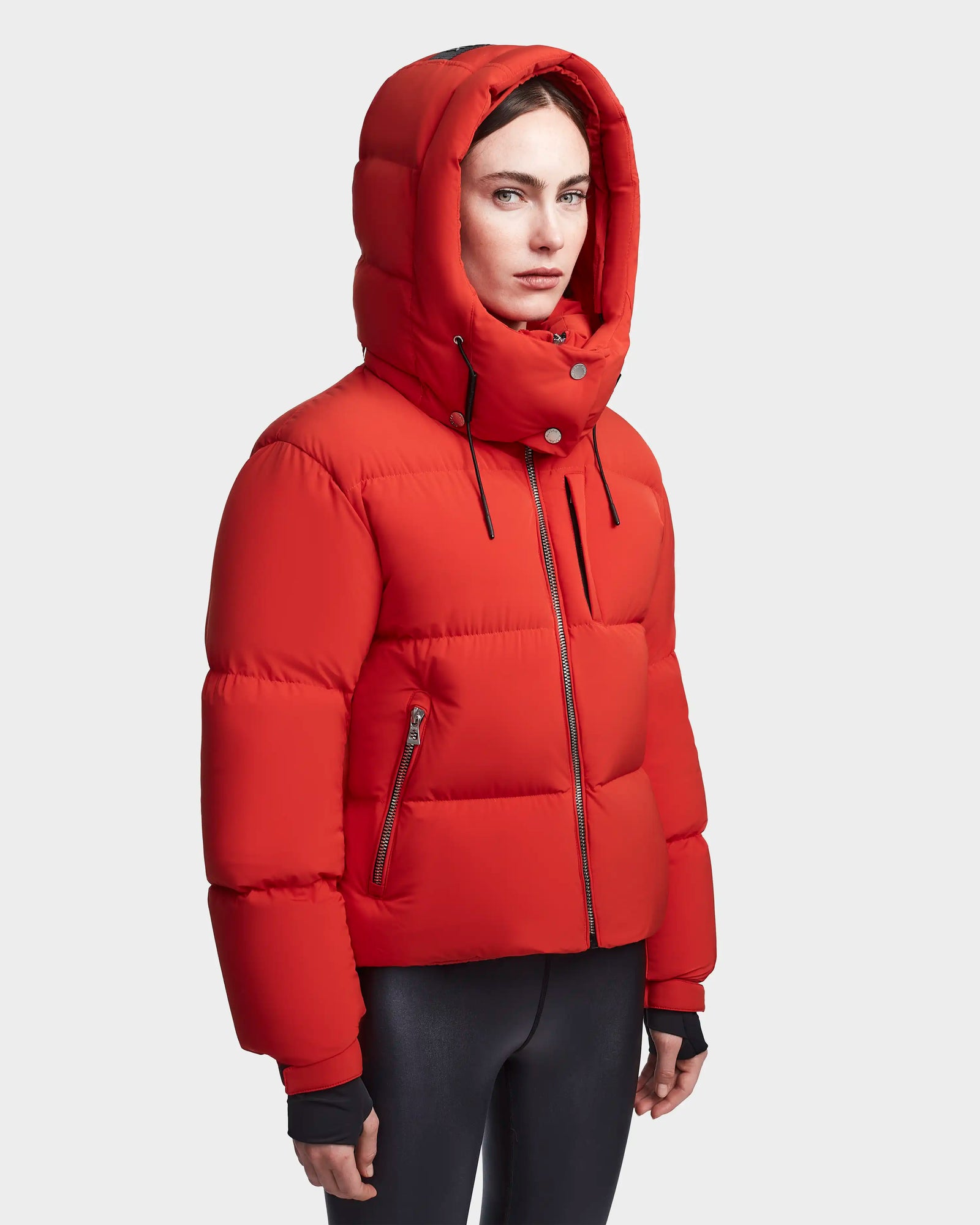 A person wearing the Rudsak MAVE M - 8123572 Orange womens down puffer jacket with a hood and black zipper, paired with black pants, stands against a plain light gray background and looks slightly to the side.