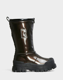 The KRISTAL HI - METALLIC CHOCOLATE by Rudsak is a shiny, dark brown high-gloss winter boot with chunky grip soles and a large R on the side, shown against a plain light grey background.