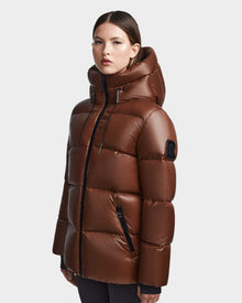 A woman stands against a light gray background wearing the Rudsak RITA S - L123527 Cohiba, a shiny brown weather-resistant puffer jacket with a high hood, black zippers, and an upper sleeve patch.