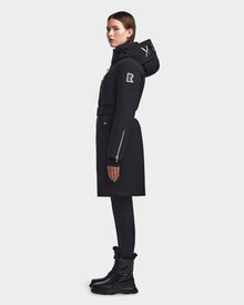 A woman in profile wears the RUDSAK MILENA - 8122536 BLACK women’s winter coat with a hood and belted waist, paired with black leggings and lace-up boots. The coat has white logo patches on the sleeve and hood. Background is plain white.