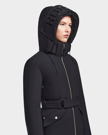 A person models the MILENA - 8122536 BLACK women’s winter coat by RUDSAK, featuring a belted waist, metallic zipper, and a long down parka hood with bold RUDSAK lettering for a chic look.
