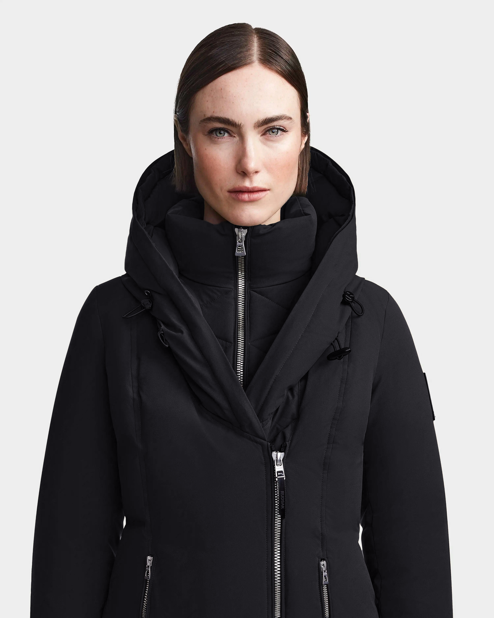 A woman with straight brown hair, light skin, and blue eyes wears the Rudsak MAIA M - 8123582 Black womens down parka, a hooded winter jacket with a high collar and double zippers, standing against a plain light background.