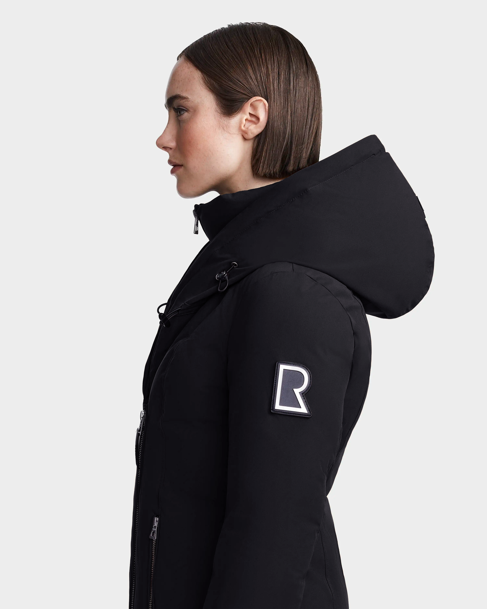 A woman with straight brown hair wears the Rudsak MAIA M - 8123582 Black winter jacket, featuring a prominent R patch on the sleeve, as she stands in profile against a plain light background.