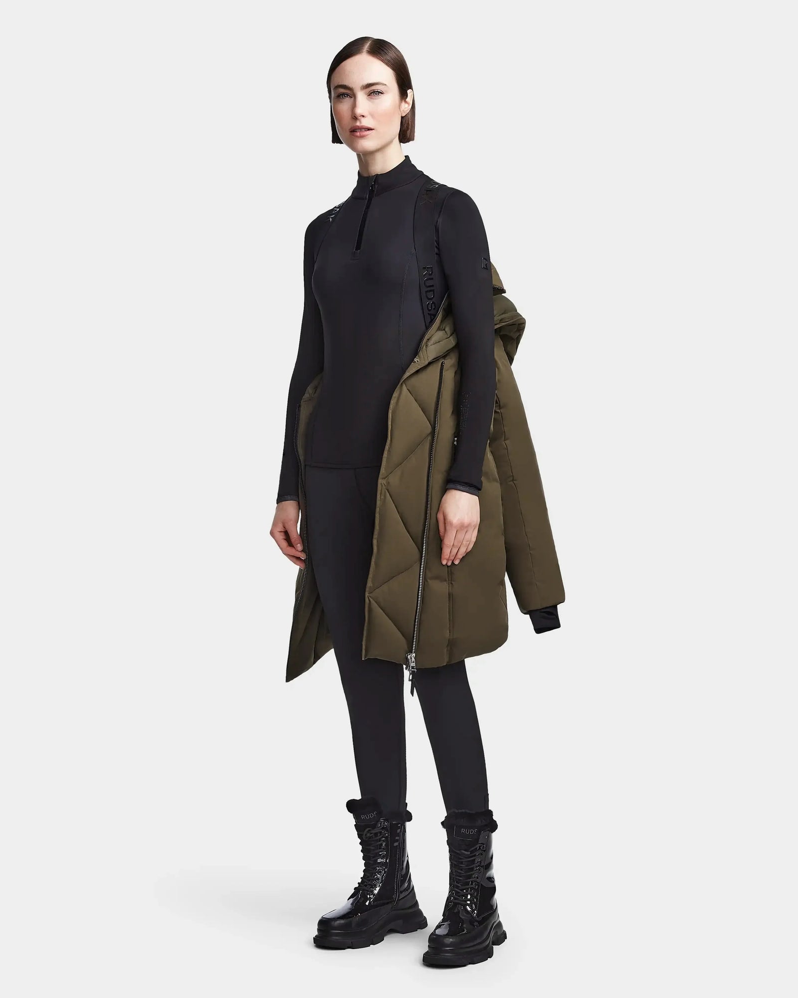 A woman poses in black fitted activewear and lace-up boots, with a Rudsak MAIA M - 8123582 Army winter jacket draped over one shoulder, set against a plain white background.