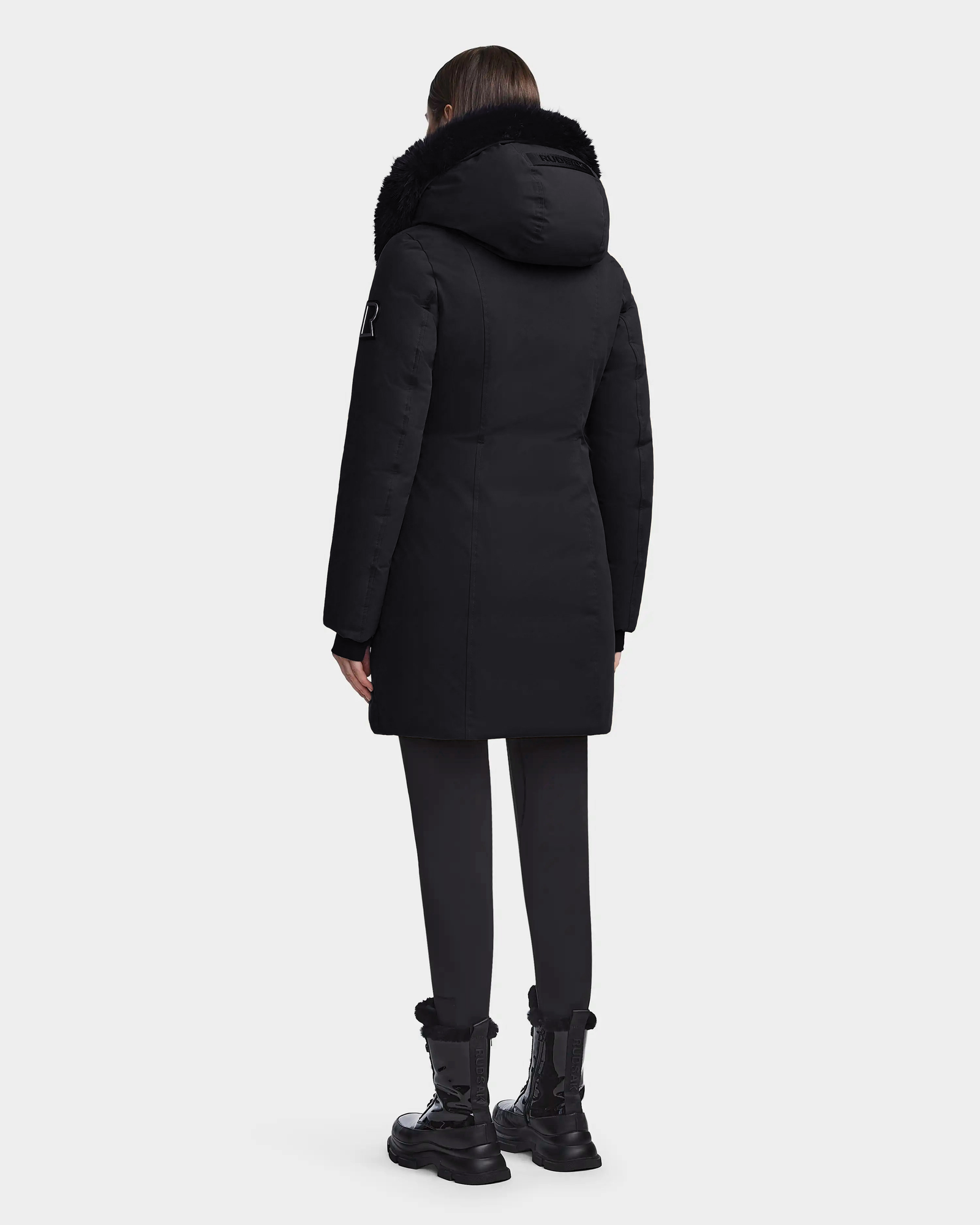 A person wearing the Rudsak MAIA M - 8123582 Black womens down parka with a faux fur hood, black pants, and black boots stands facing away from the camera against a plain white background.