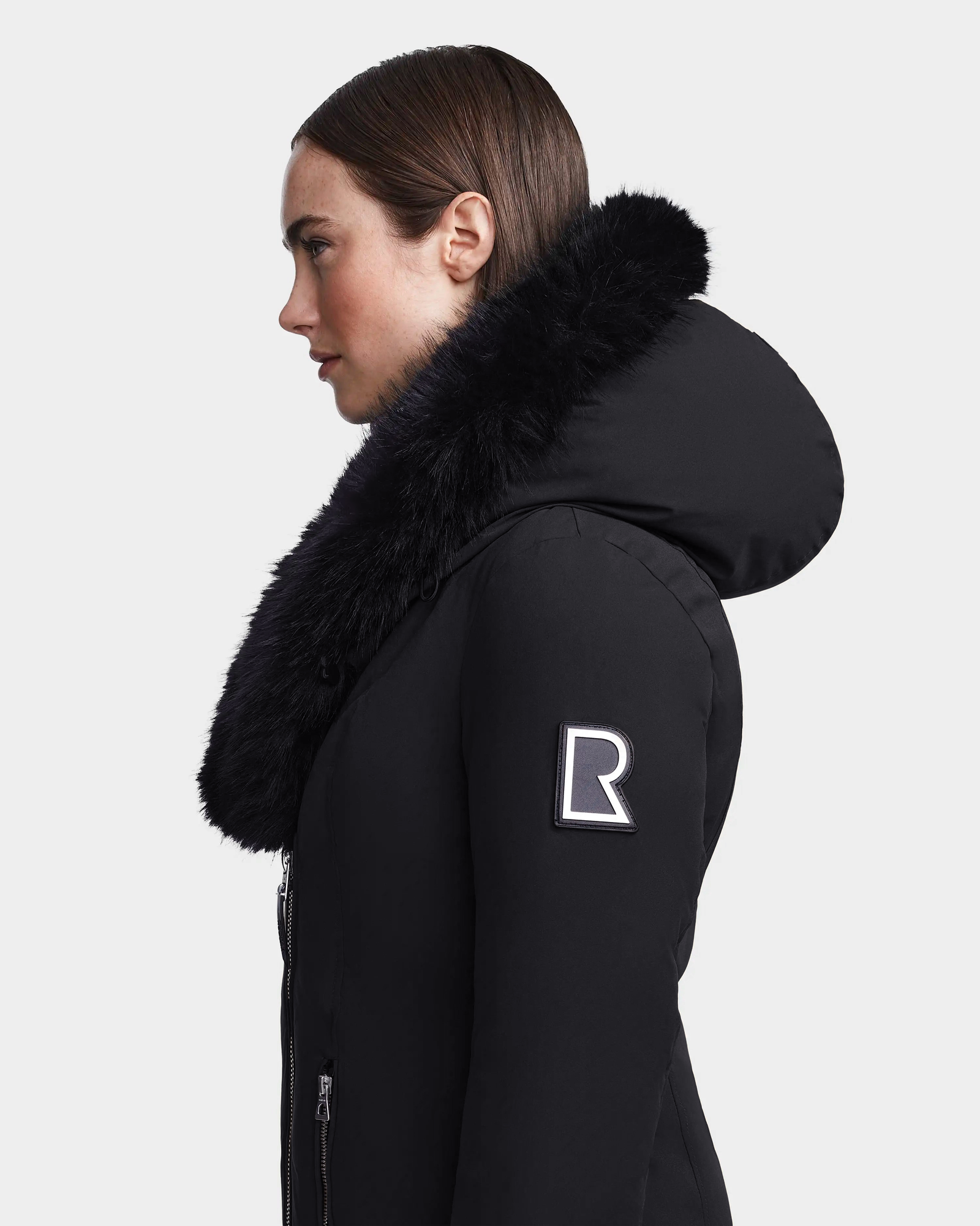 A woman in profile wears the Rudsak MAIA M - 8123582 Black winter jacket with a faux fur hood and an R patch on the sleeve. Her straight hair is tucked behind her ear, set against a plain white background.