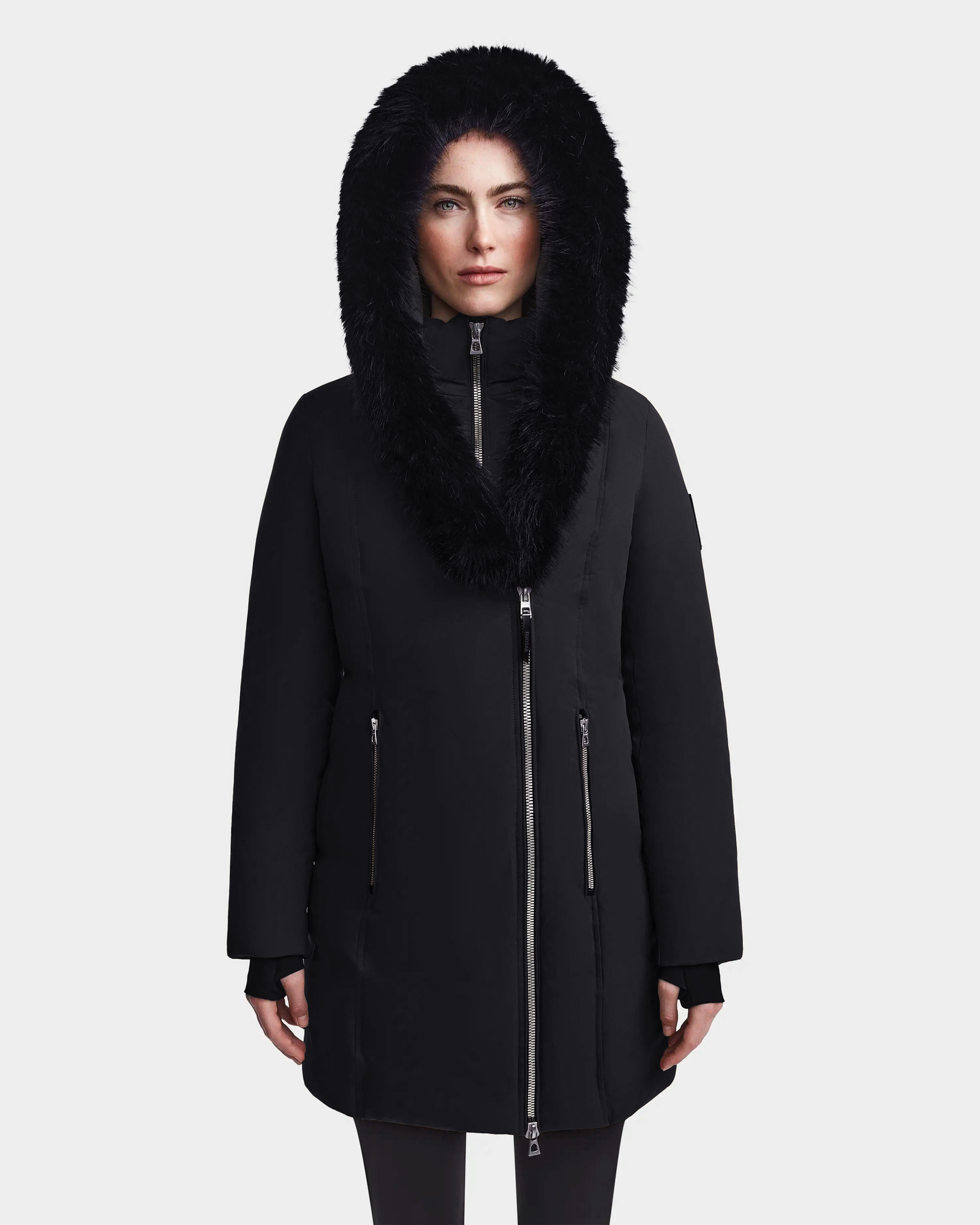 A woman models the Rudsak MAIA M - 8123582 Black down parka with a zip-up front, zippered pockets, and a black faux fur-lined hood. She faces forward against a plain white background with her hands by her sides.