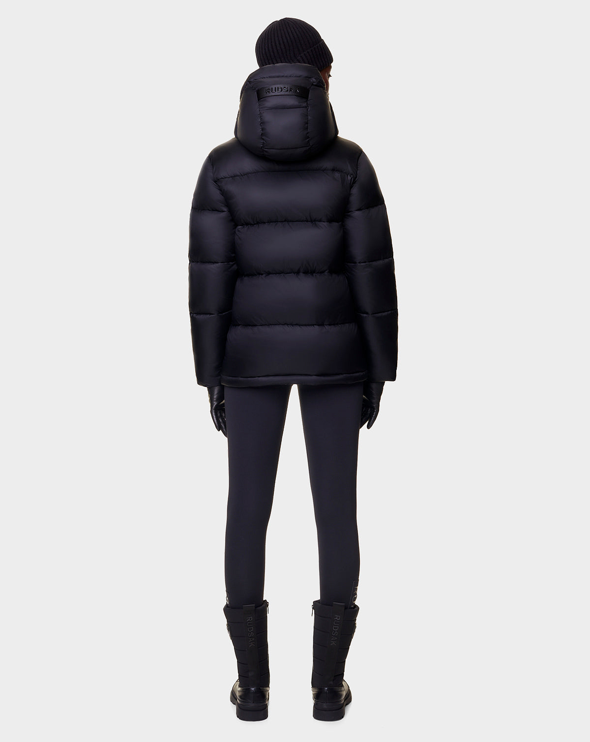 A person wearing the Rudsak EVIE - L122517 BLACK puffer, paired with black pants, gloves, boots, and a beanie, stands facing away against a plain light background.