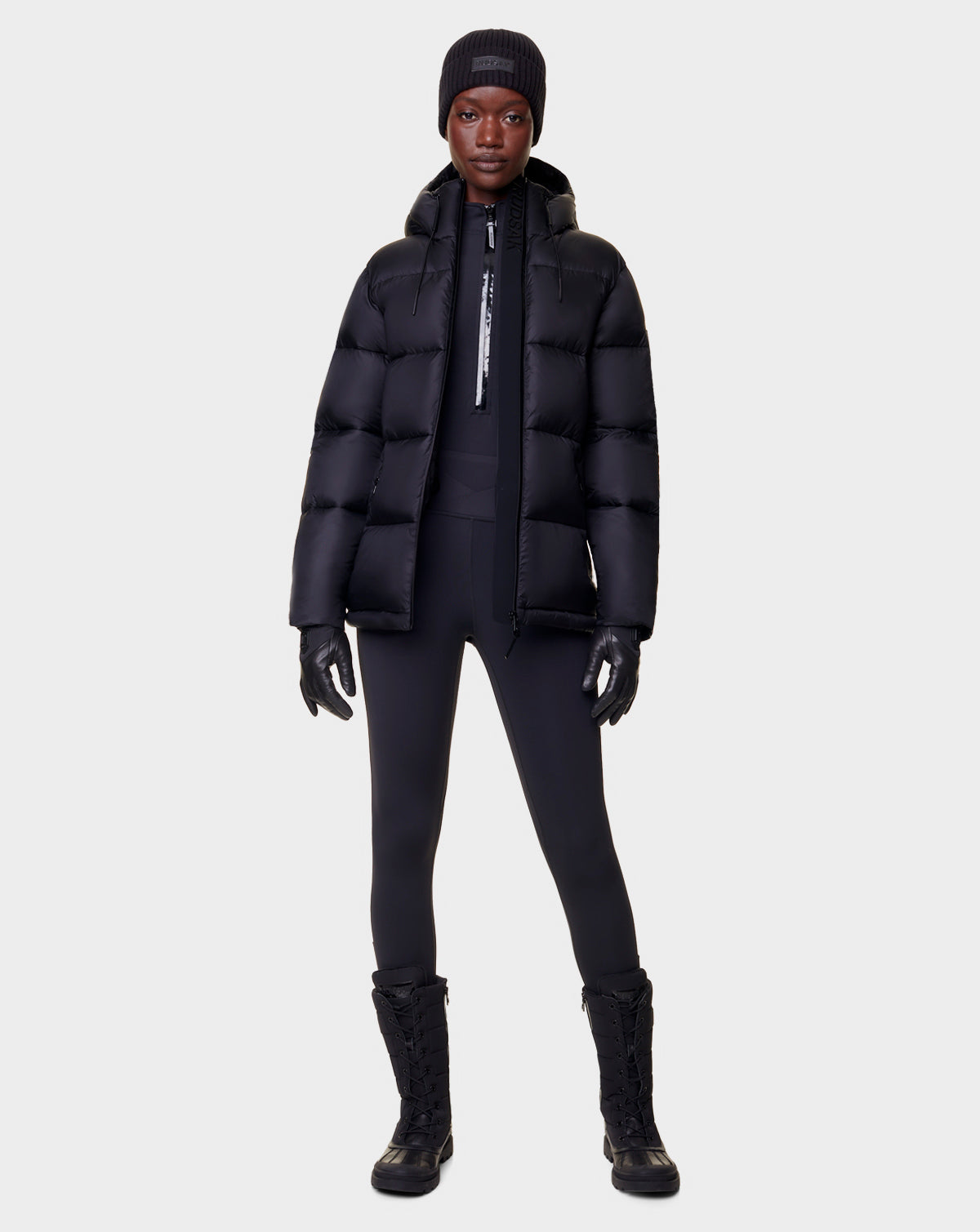 A person faces forward in a Rudsak EVIE - L122517 BLACK womens RDS-certified down puffer jacket, styled with black gloves, leggings, lace-up boots, and a beanie against a plain light gray background.