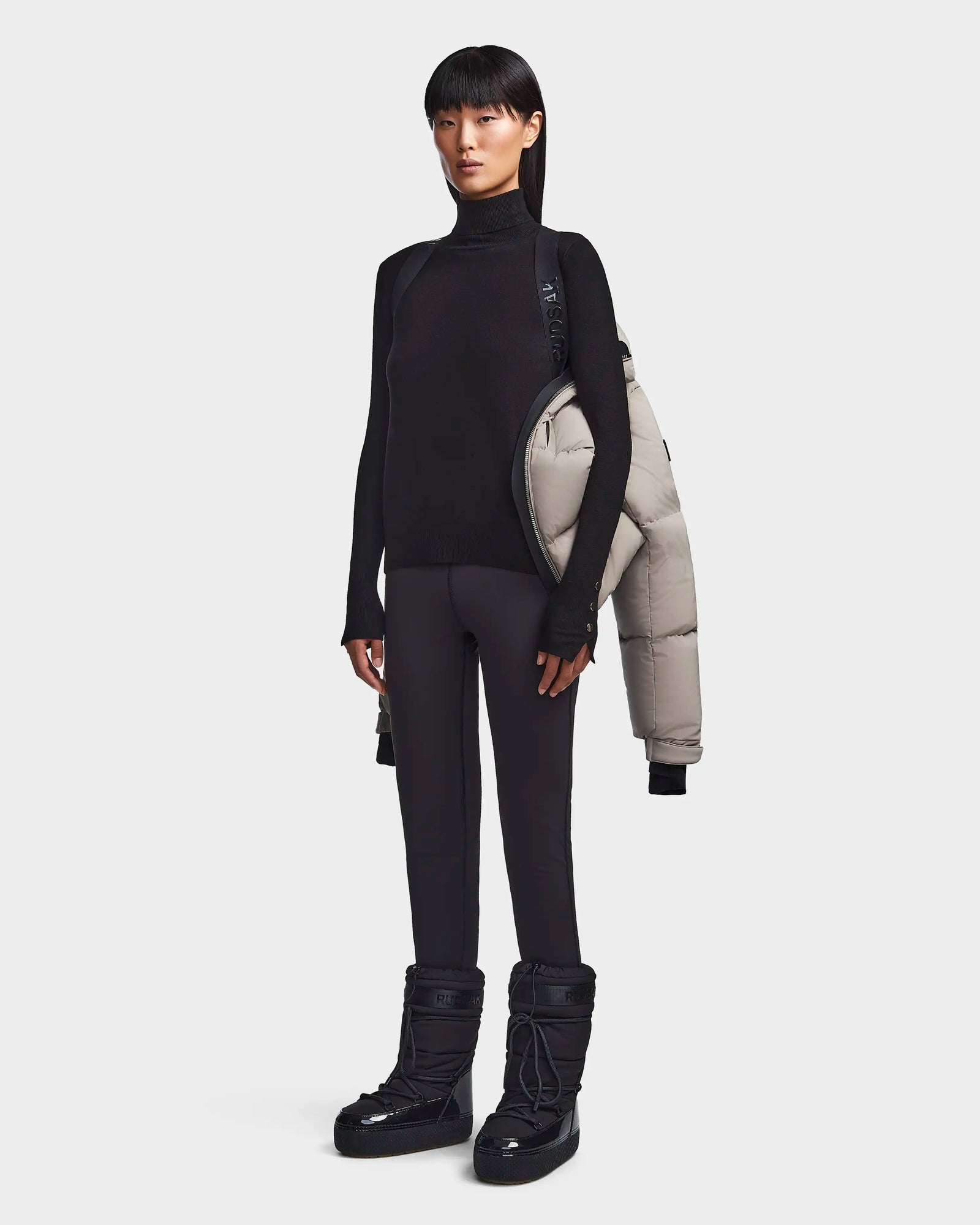 A woman with straight dark hair, dressed in black, holds the Rudsak MAVE M - 8123572 Cement women’s down puffer over her left shoulder while standing against a plain white background.